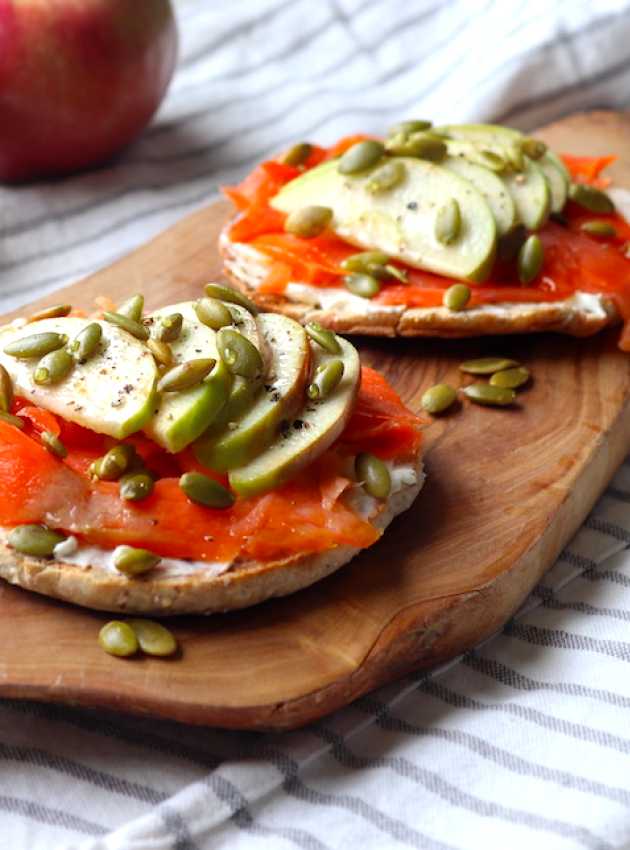 Bagels garnis de saumon fumé, tranches de pomme, graines de citrouille et fromage à la crème sur une planche en bois.