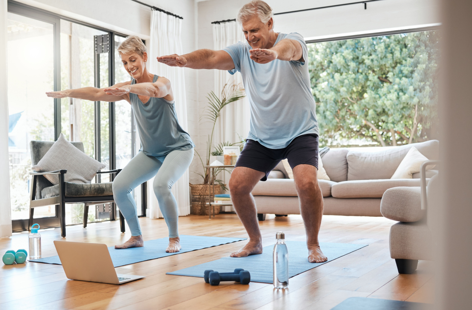  seniors watch a computer as they bend their knees and stretch their arms on yoga mats at home.