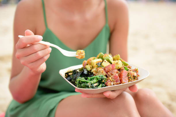 A woman enjoying a healthy Hawaiian food bowl by the beach, with fresh ingredients like poke, limu, and tropical fruit.