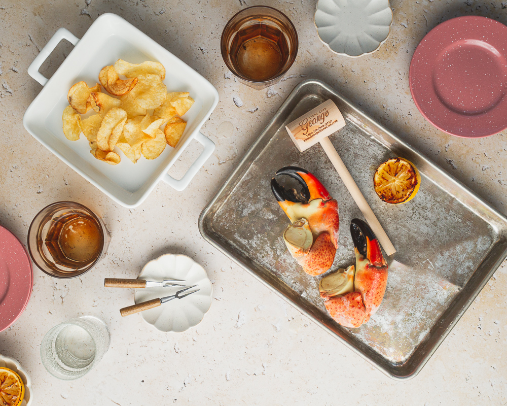 Stone crab claws on a metal tray served with wooden mallet accompanied by a side of potato chips, grilled lemon, and drinks