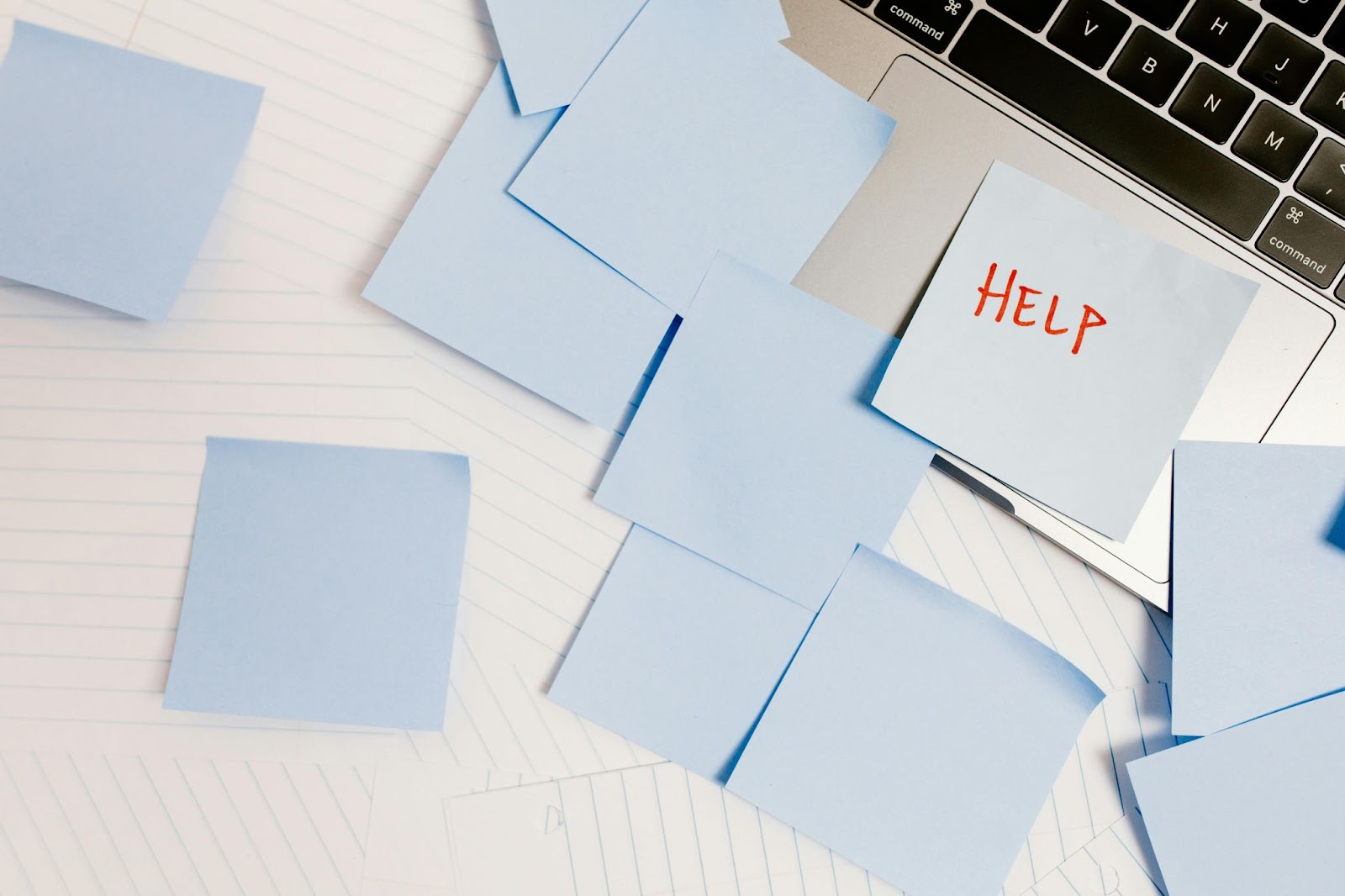 Light blue Post-it notes litter a desk covered with lined paper and an open laptop. One Post-it note reads, in red capital letters: 'HELP'.