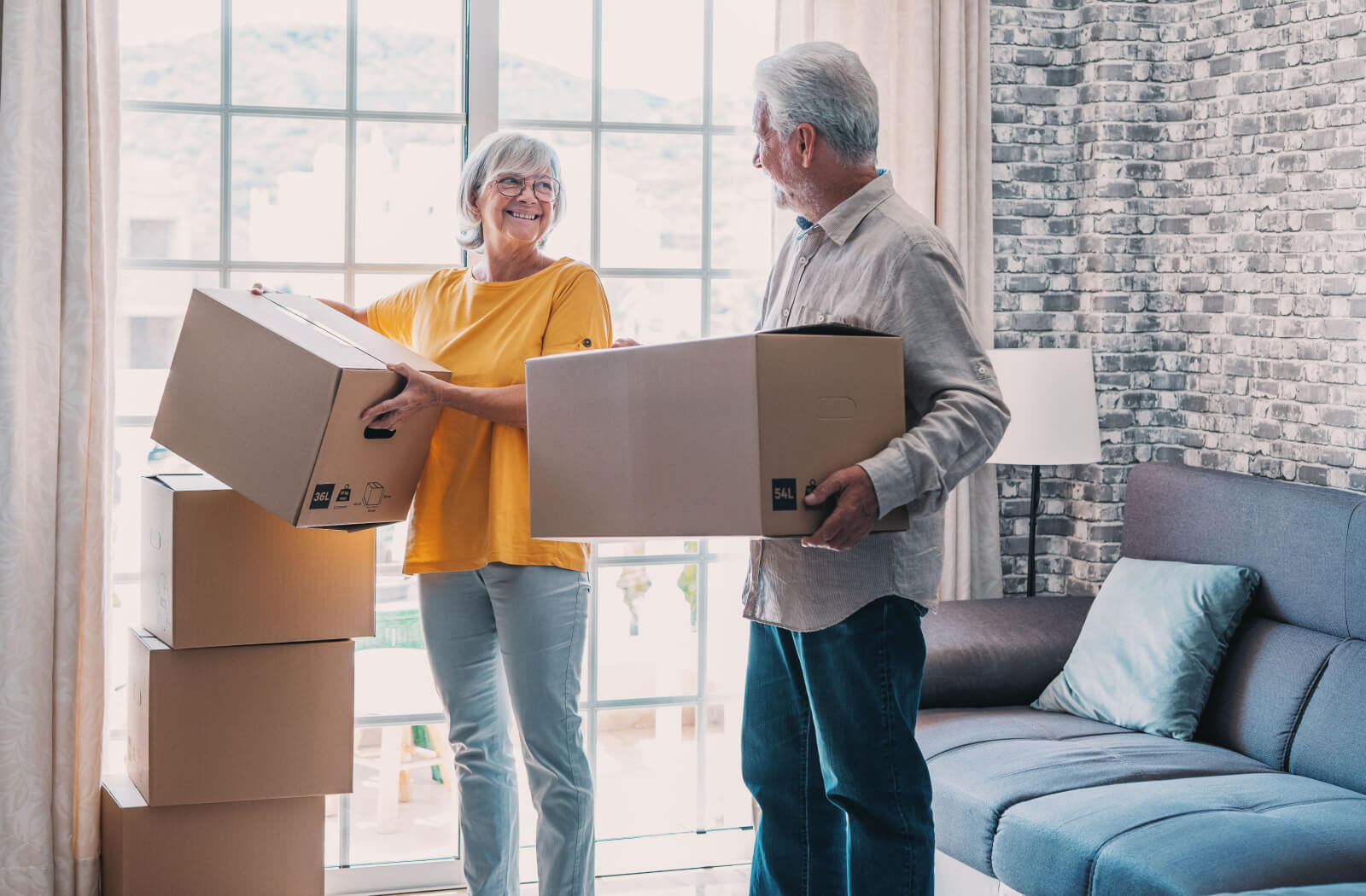 An older married couple stacking moving boxes in front of a sunlit window, smiling at one another and happy.
