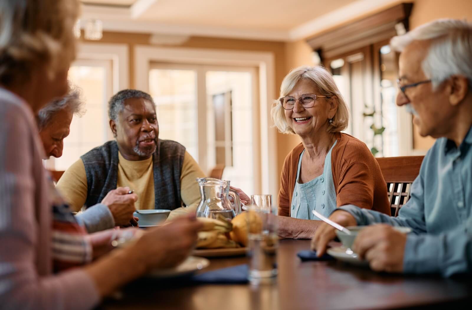  A group of seniors smiling and laughing over breakfast in a communal dining room lit by soft yellow light