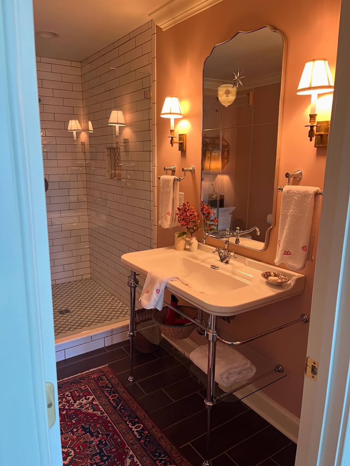 A bathroom with lit wall sconces features blush pink walls, a chrome-framed sink, a large mirror 