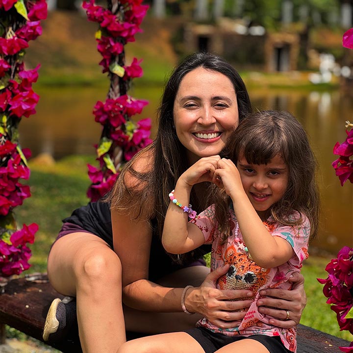 Mãe e filha sorrindo juntas ao ar livre, cercadas por flores rosa e fundo de lago e árvores, em uma cena de alegria e conexão familiar.