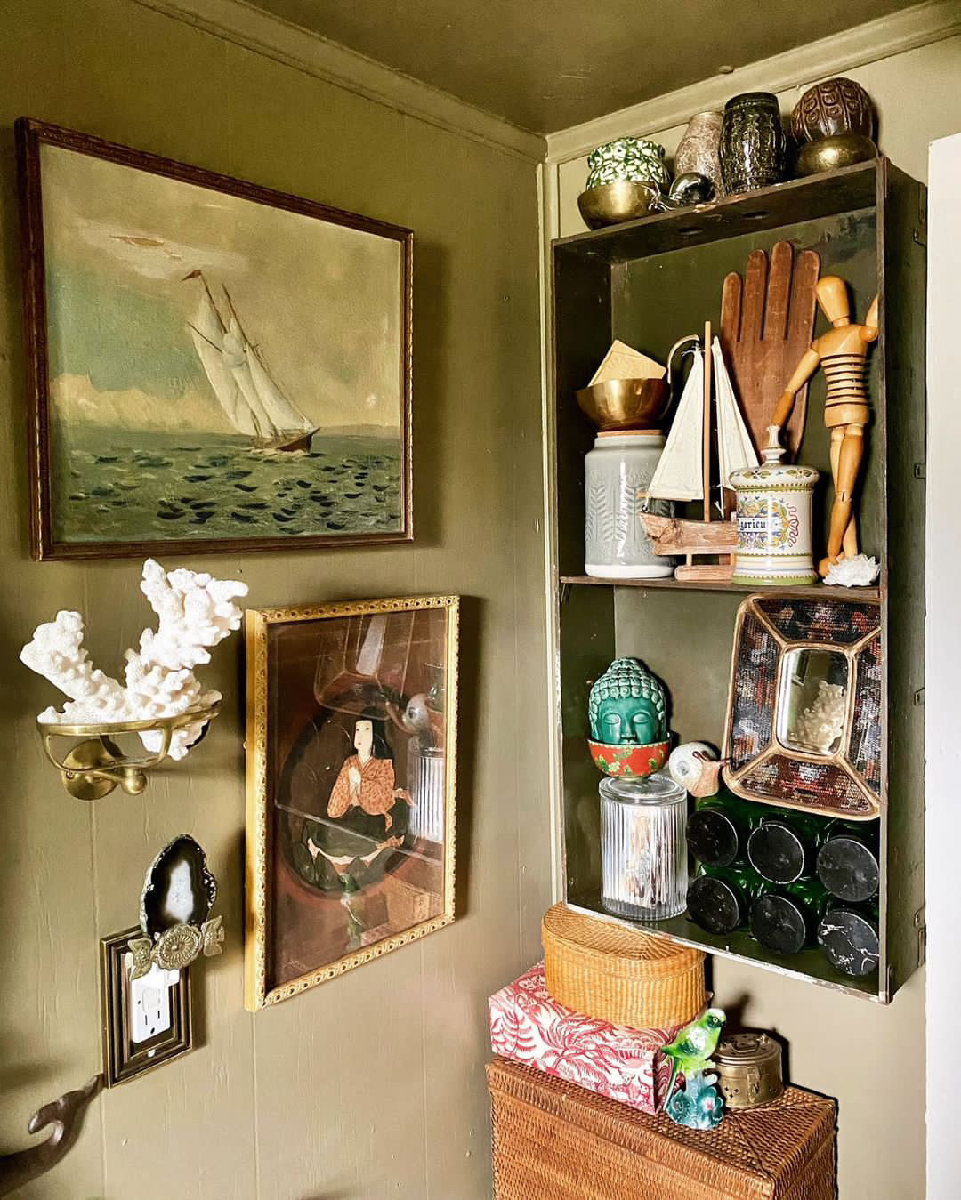 A corner of the room features 2 framed artworks, a coral piece, and a vintage mirror on a muted olive-green wall. One is a painting of a sailboat at the sea, and the other is a portrait of a seated lady. A small shelf holds an assortment of decorative objects including a wooden mannequin, a Buddha head, and various vases and bowls. Below the shelf is a wicker basket.