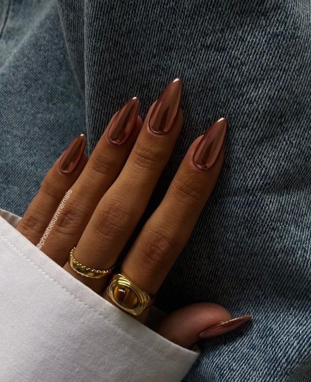 Close-up of a hand with glossy, long, almond-shaped brown nails and gold rings against denim fabric.

