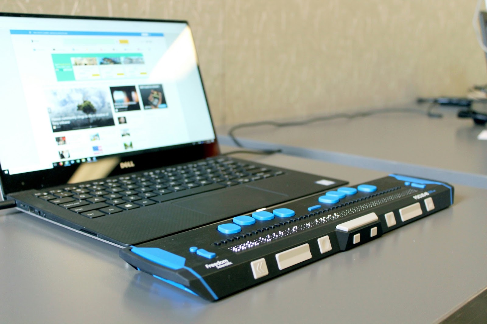 A laptop connected to a refreshable braille display with blue and white buttons, sitting on a desk. The laptop screen shows a website interface.