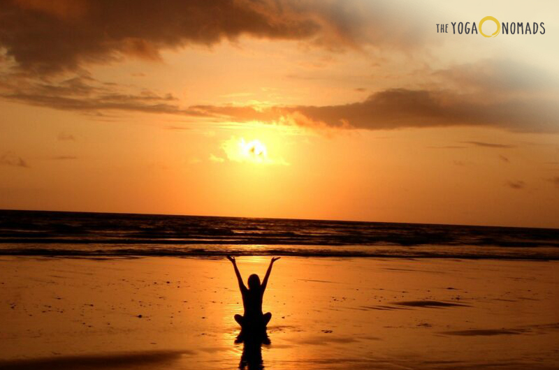 crown chakra: A beach at sunset with a person in the distance raising their arms towards the sky, creating a reflection on the wet sand. The sky is adorned with warm hues of orange and yellow around the sun, transitioning to darker shades towards the edges of the image.