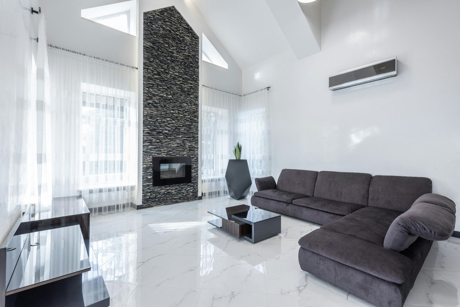 A modern living room with a sleek gray couch, a stone-textured fireplace, and a mounted AC unit on a clean white wall