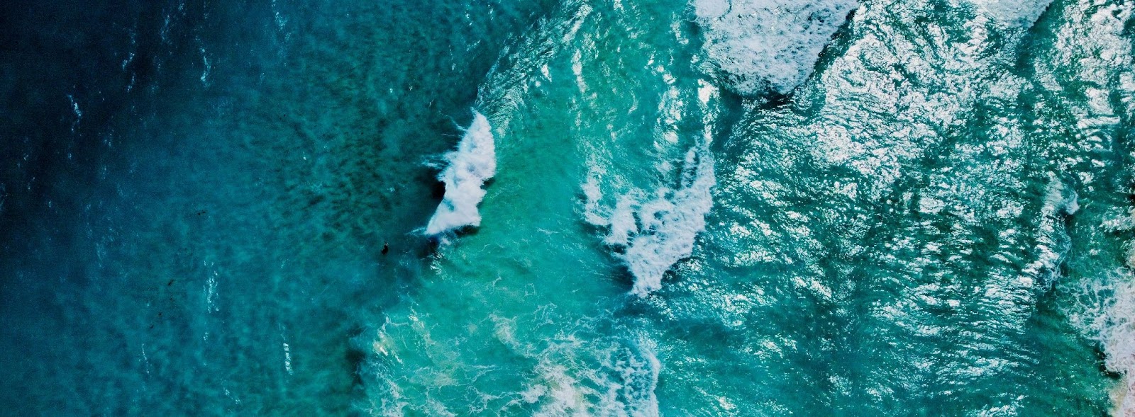Vista aérea de ondas de Cancún quebrando em um mar azul-turquesa cristalino, com diferentes tons de azul destacando a profundidade e a beleza da água.