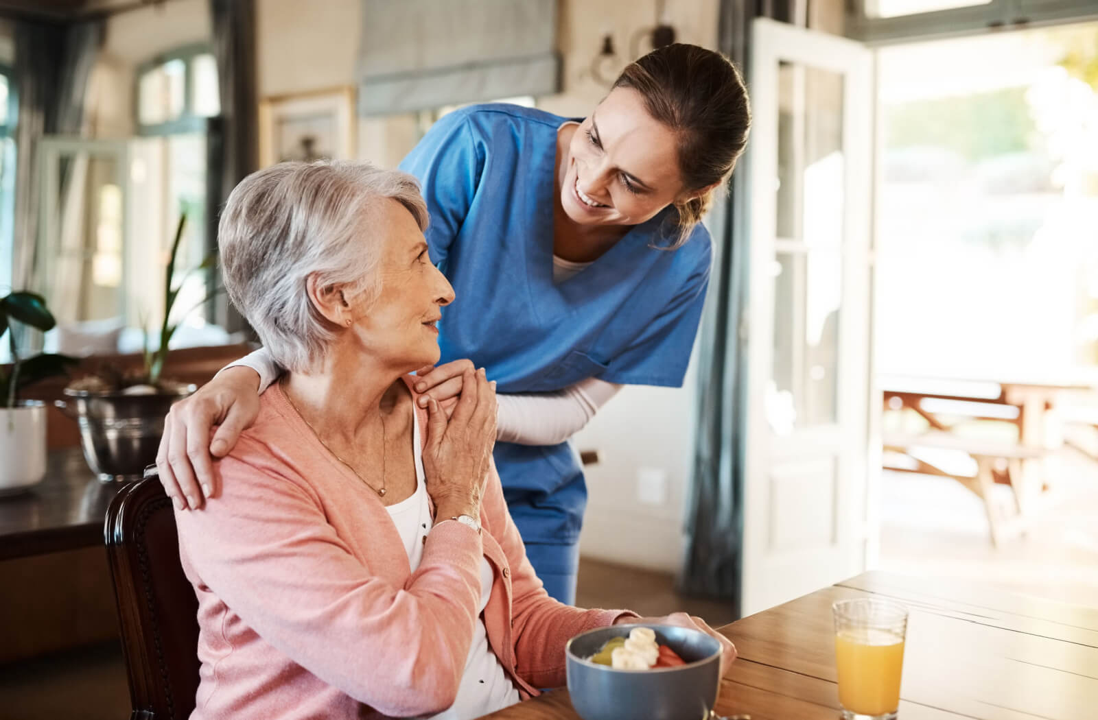 A caregiver with her arm around a senior woman smiling at each other during breakfast in memory care