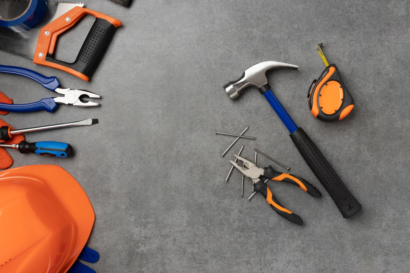 A variety of construction tools for Garage Doors neatly arranged on a gray surface, including a hammer, pliers, a tape measure, nails, screwdrivers, a saw, and an orange safety helmet, showcasing essential equipment for construction or repair work