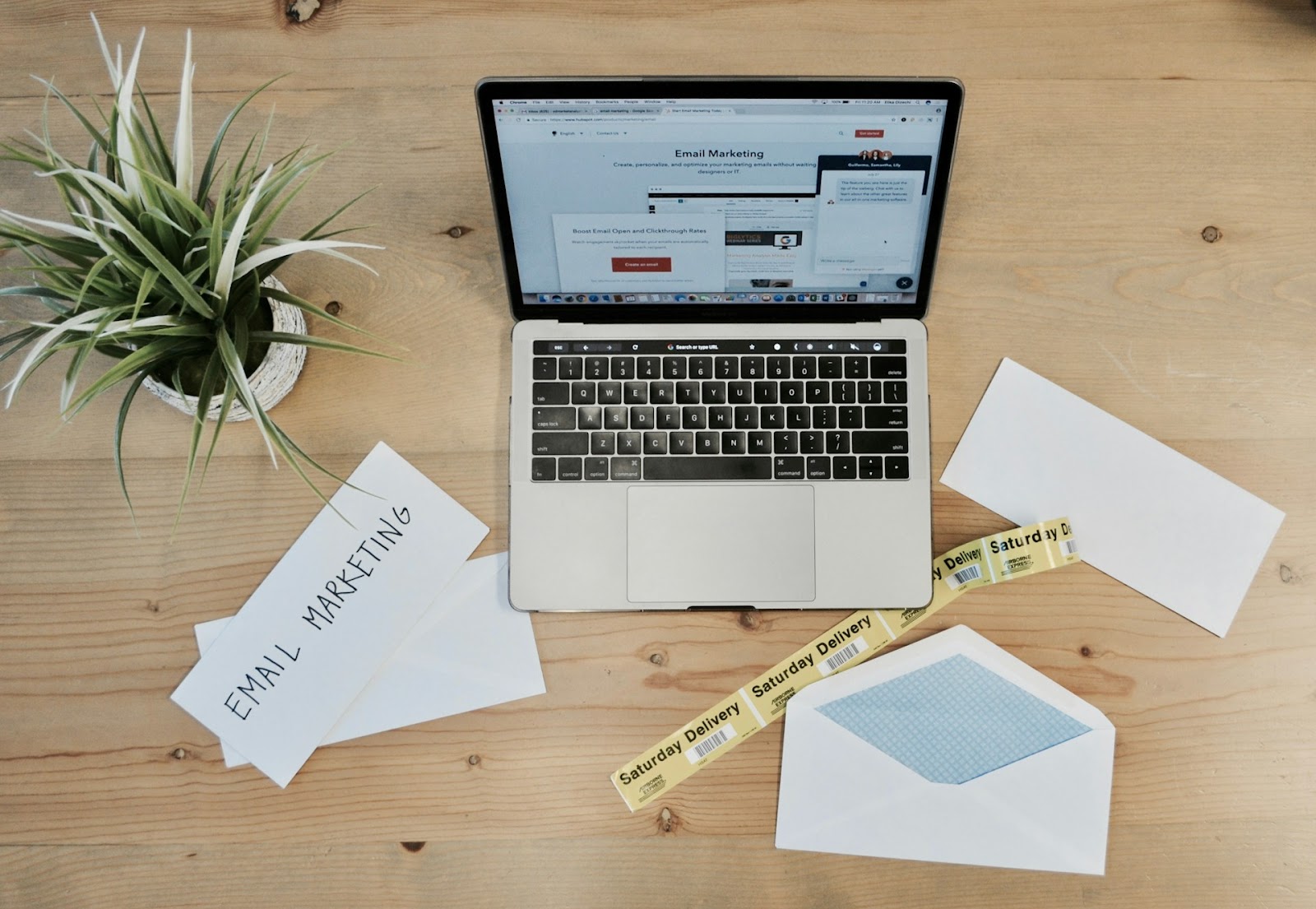 A laptop displaying an email marketing platform on a desk, surrounded by envelopes, a plant, and 'Saturday Delivery' labels, emphasizing the concept of email marketing as marketing automation best practices