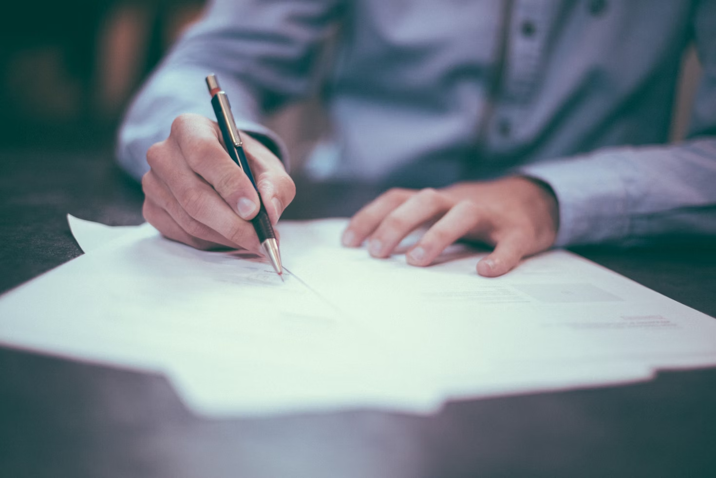 A person writing on documents with a pen, wearing a long-sleeved shirt, with papers spread out on a desk