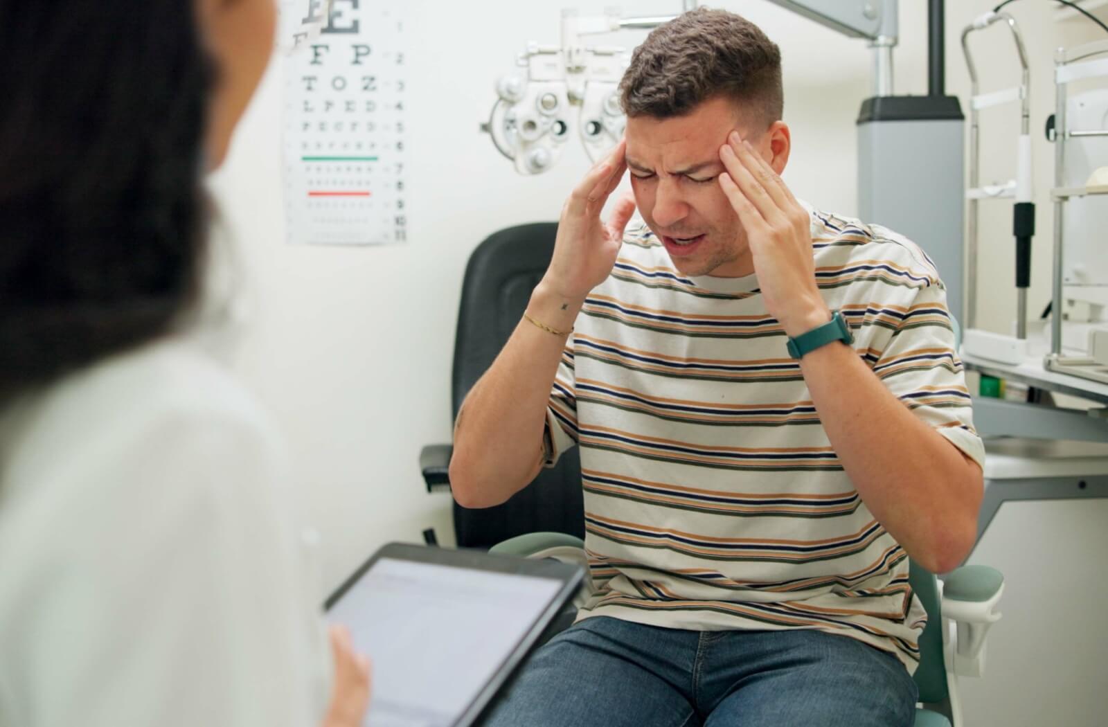 a patient rubs their temples while explaining symptoms of dry eyes and headaches to their optometrist during a consultation