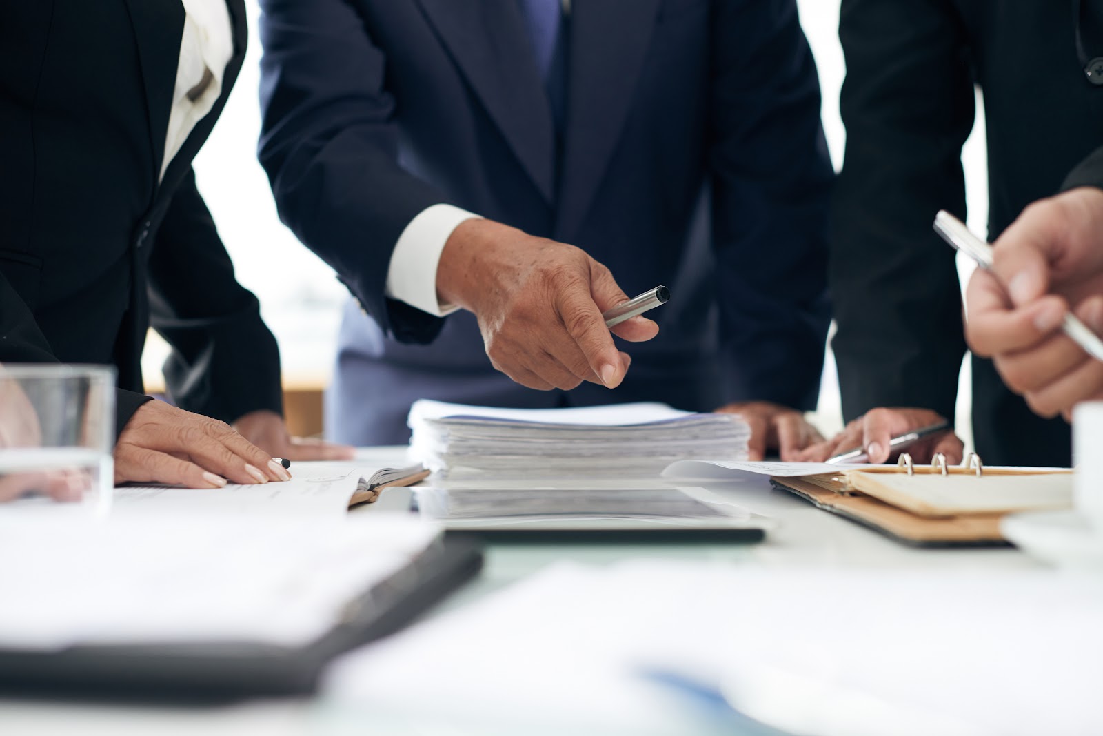 A group of business professionals gathered around a table, reviewing documents and pointing with pens.