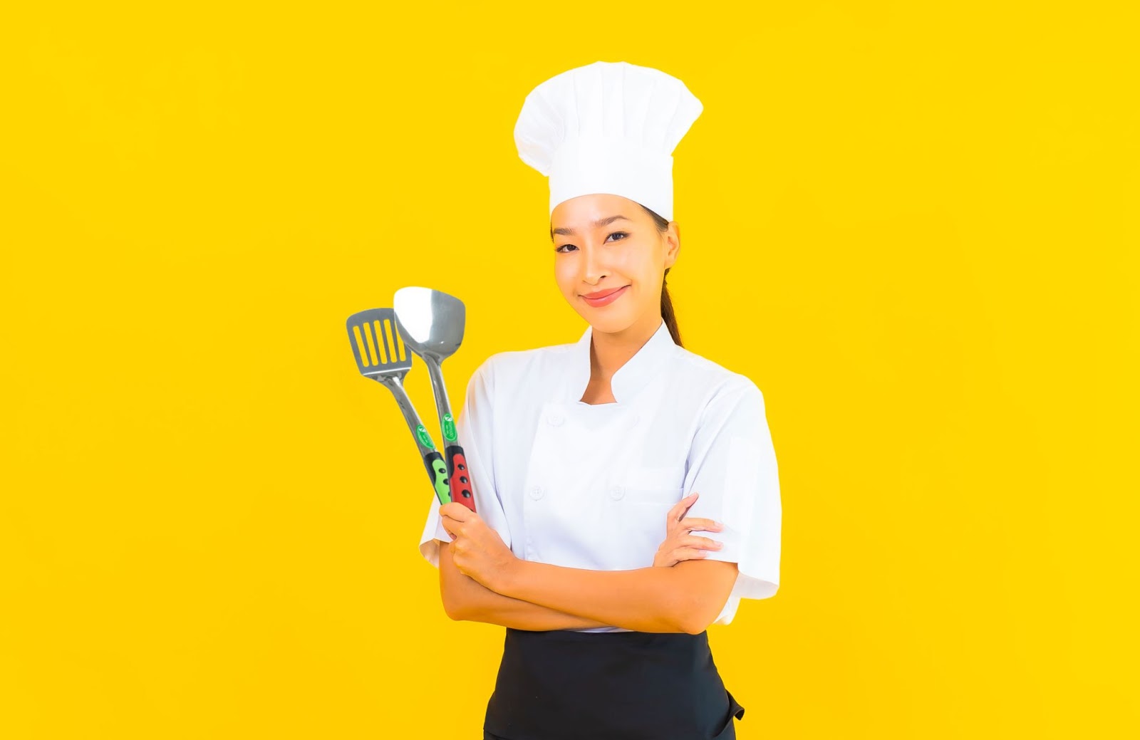 Koki wanita Asia memegang spatula dengan latar belakang kuning cerah, siap memasak dengan semangat.
