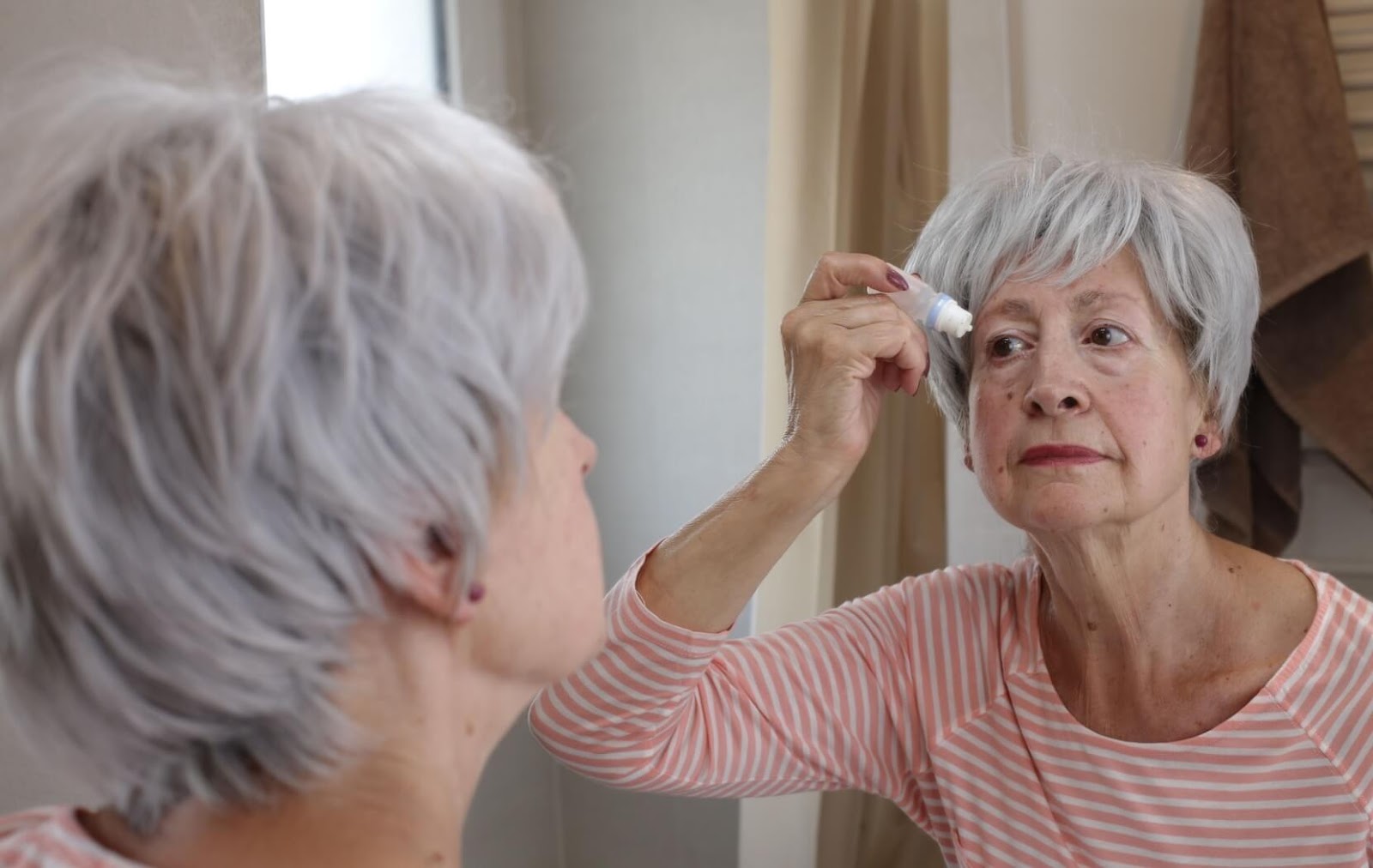 A senior looks in their bathroom mirror while they apply eye drops to help relieve dry eye symptoms.