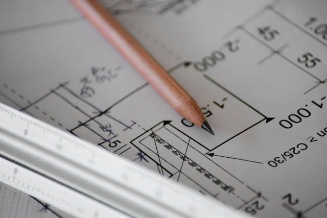 A close-up of architectural plans with a pencil and ruler resting on top. The plans, intricately detailed with measurements and technical drawings, incorporate interior design elements that focus on ergonomic solutions to ensure comfort and efficiency in the space.