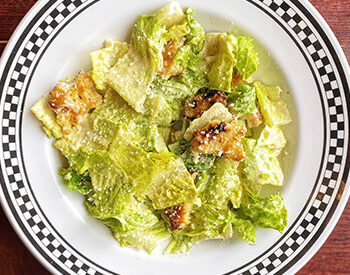 A classic Caesar salad with romaine lettuce, crunchy croutons, and grated cheese, served in a white plate with a black checkered rim.