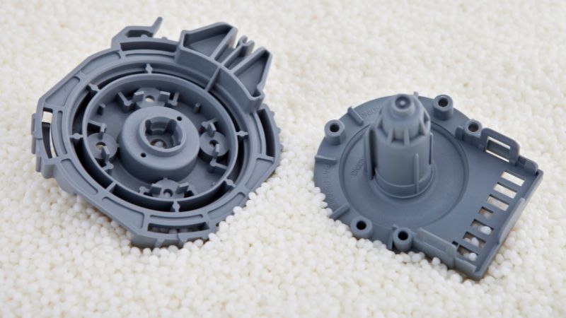 Two gray plastic gears rest on a surface covered in small white pellets.