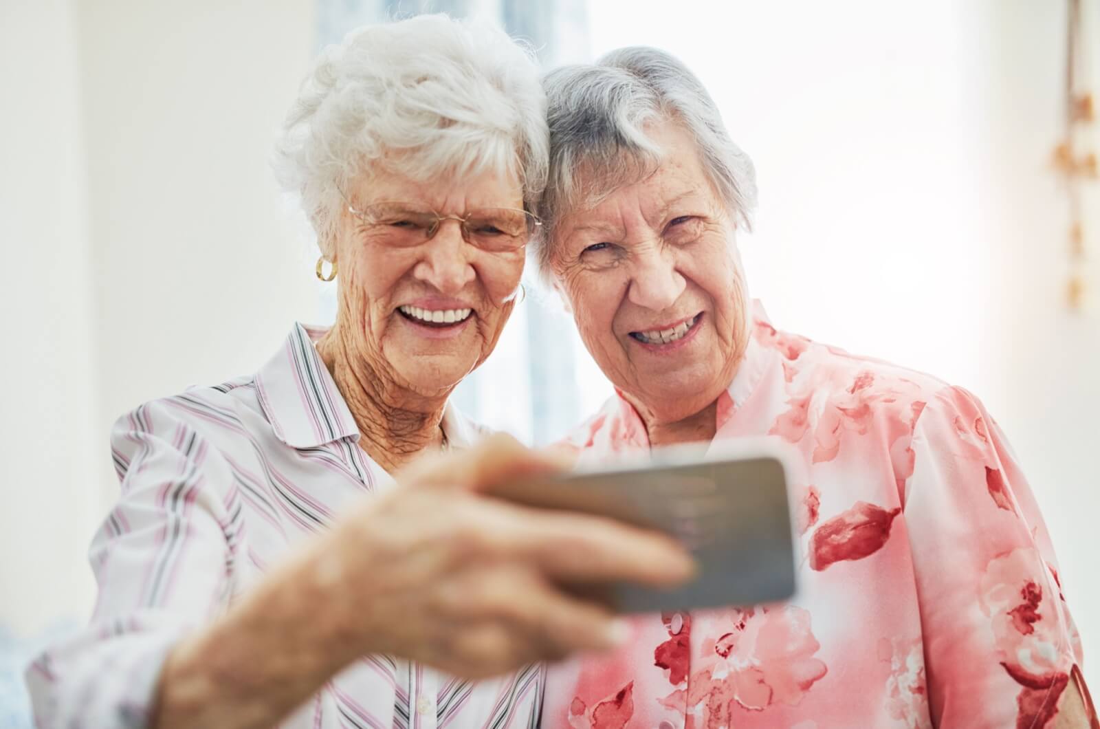 Two smiling older adults standing side-by-side with a cell phone held in front of them in one hand.