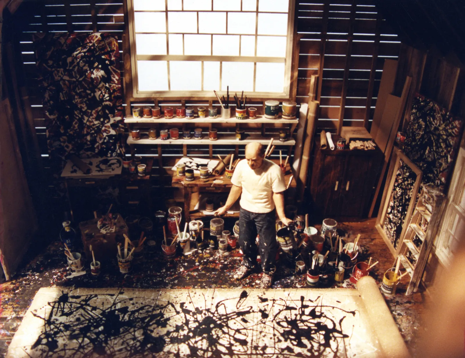 Jackson Pollock in his art studio surrounded by paint cans and brushes, creating an abstract drip painting on a large canvas, showcasing his signature action painting technique.
