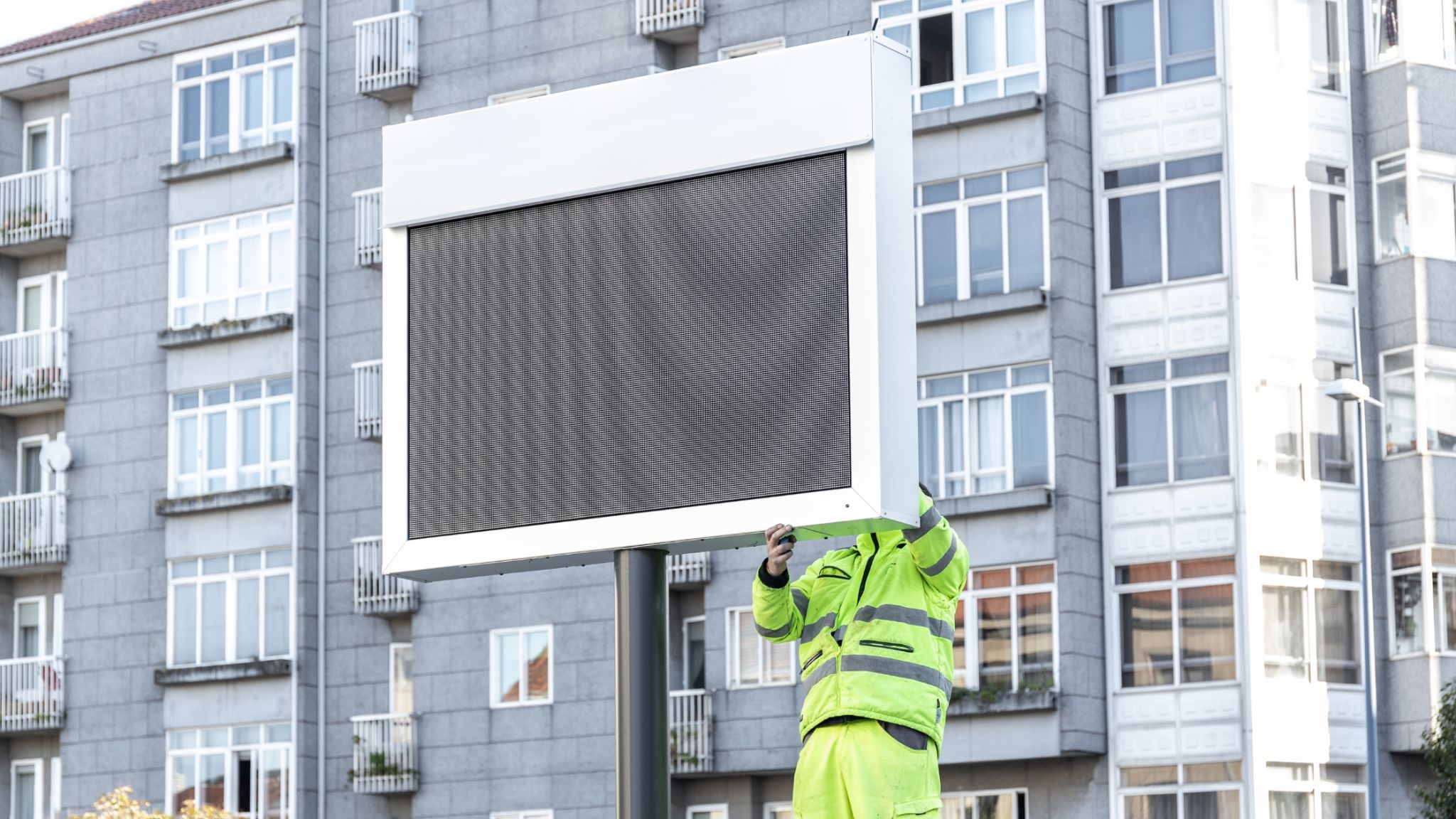 A technician in a high-visibility uniform installing an outdoor digital display in Qatar, ensuring secure setup in an urban environment with residential buildings in the background.