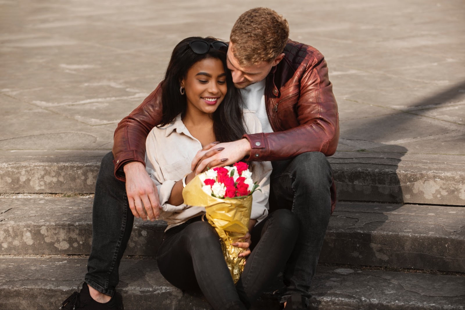 A couple sitting on stairs, where the man hugs the woman from behind. The woman is holding a bouquet of flowers in her hand and hold with her other hand the hand from her lover.