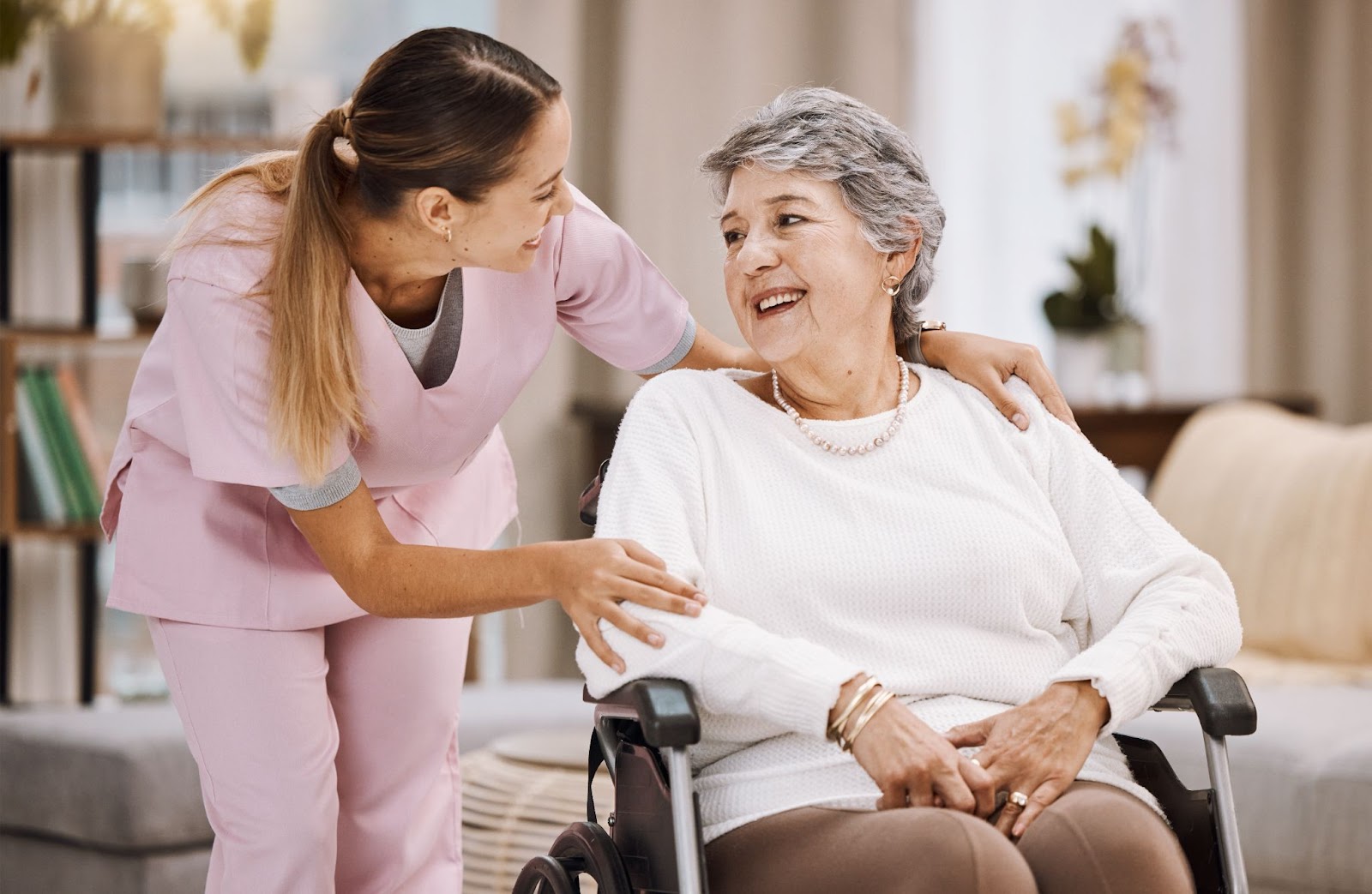 A female caregiver in an assisted living community bends down to talk to a smiling older adult resident in a wheelchair.