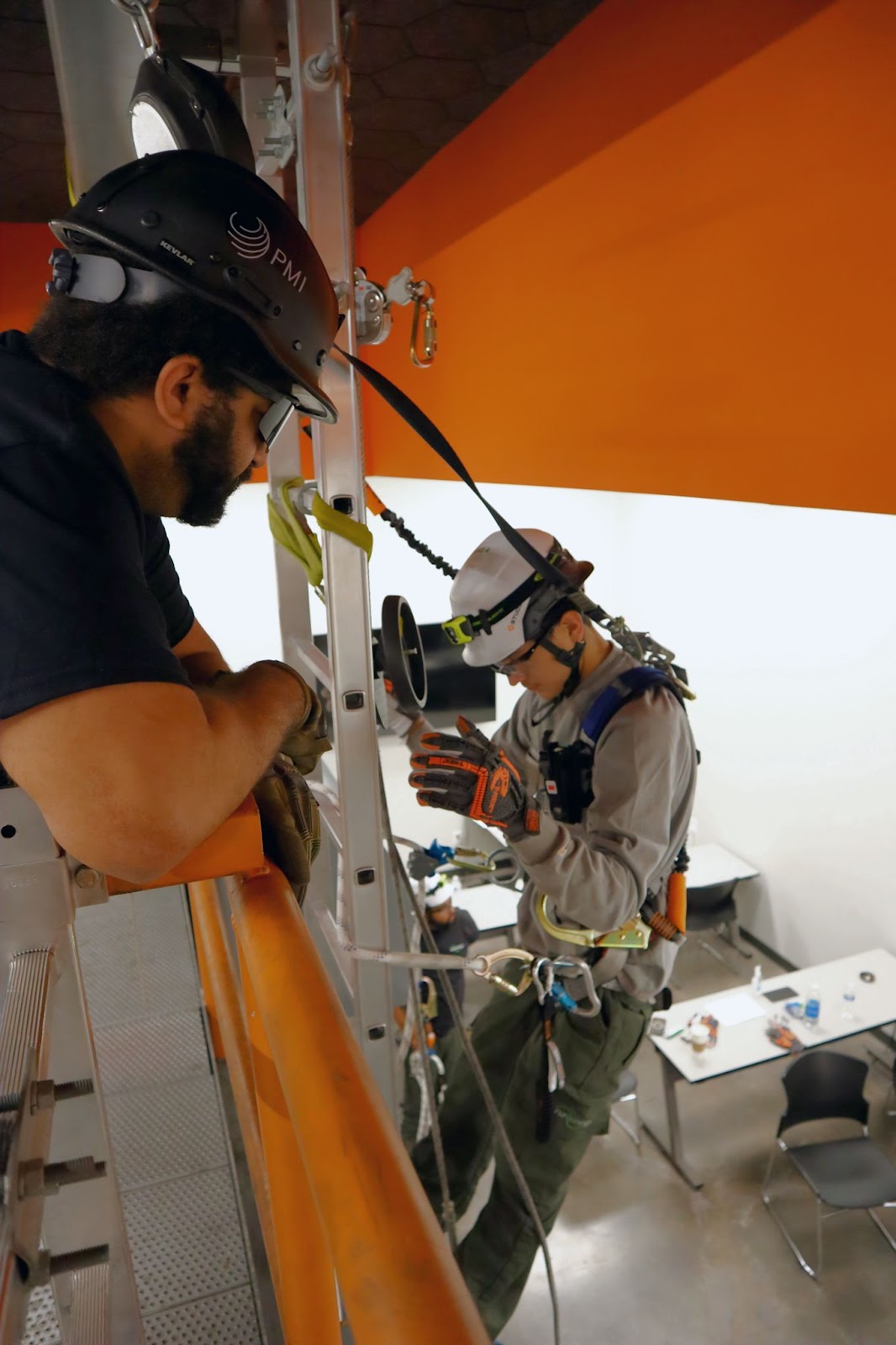 Two individuals engaged in safety training on a high platform, with one observing and the other wearing a harness and helmet while working on a secured ladder. The environment reflects a focus on technical skills and safety, with an orange railing adding a pop of color to the industrial setting.