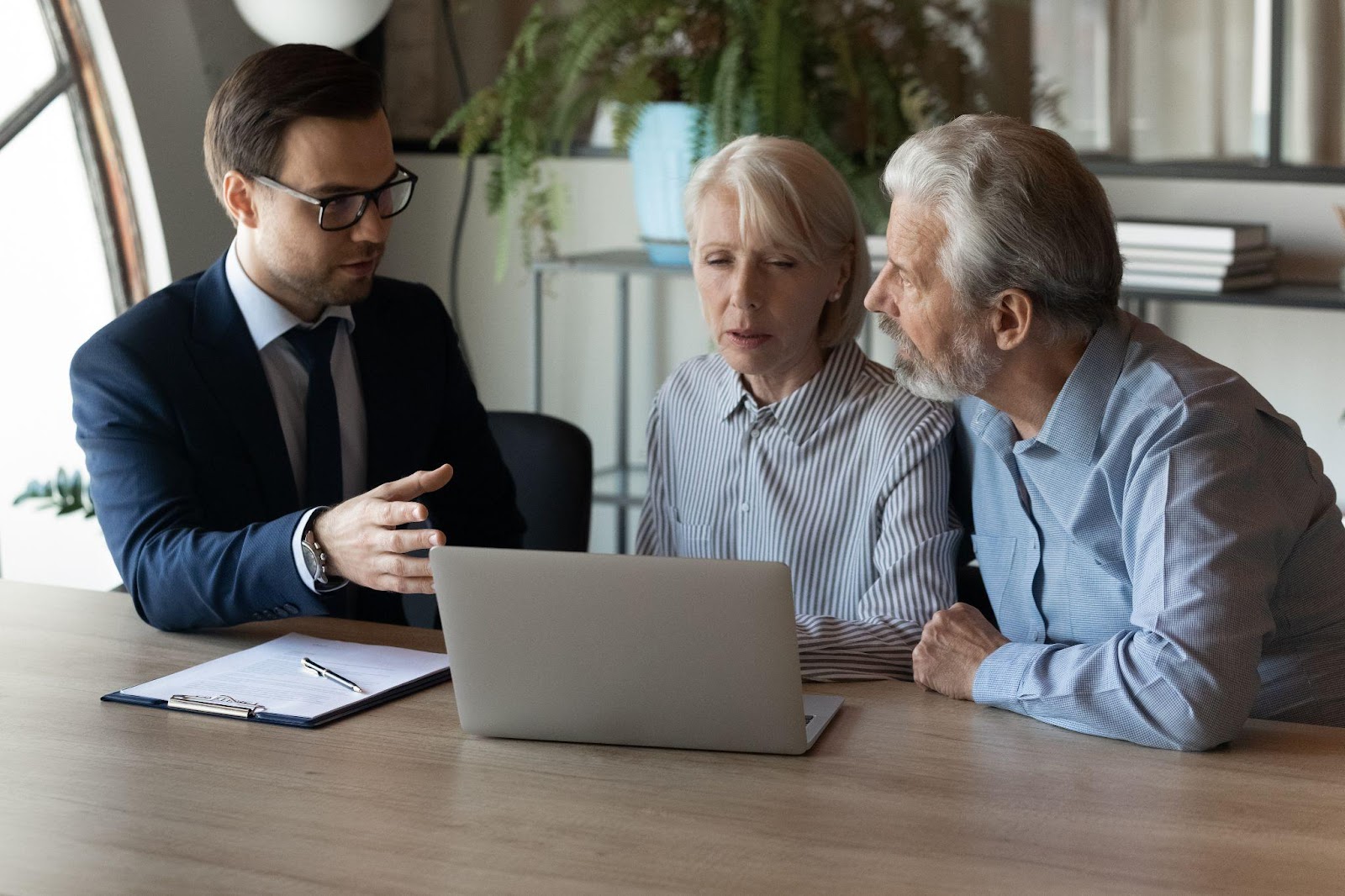 A couple sits at a conference table with a lawyer. A laptop is sitting on the table in front of them. The lawyer is gesturing at the laptop screen while explaining its contents to their clients.