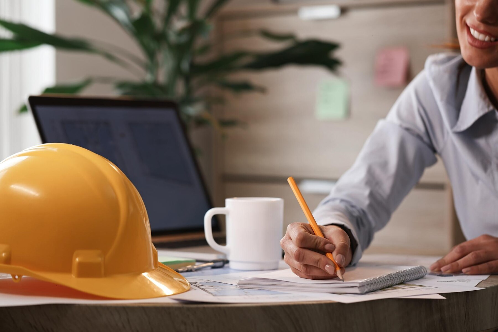 Amidst a clutter of papers, a person writes notes next to a yellow hard hat, a white mug, and a laptop. The scene, enhanced by the lively touch of a plant and filing cabinet in the background, reflects an office-like setting ripe for exploring key skills in architectural journalism.