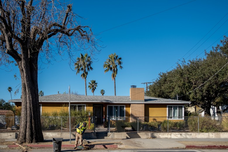 A yellow single-story house with a gray roof is visible behind a chain-link fence, set against a backdrop of a clear blue sky and tall palm trees. In the foreground, a person wearing a high-visibility vest and khaki pants is sweeping the sidewalk near a leafless tree and a trash bin. The scene is calm, with utility wires stretching across the frame.