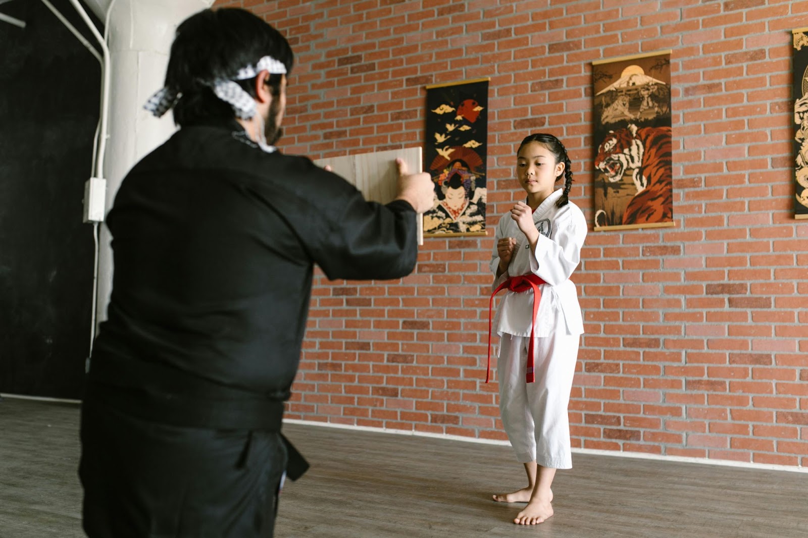 A martial arts instructor kneels on one knee, holding out a piece of paper toward a young student