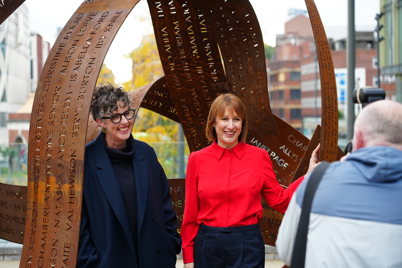 Two women stand in a sculpture of metal ribbons, being photographed by a man