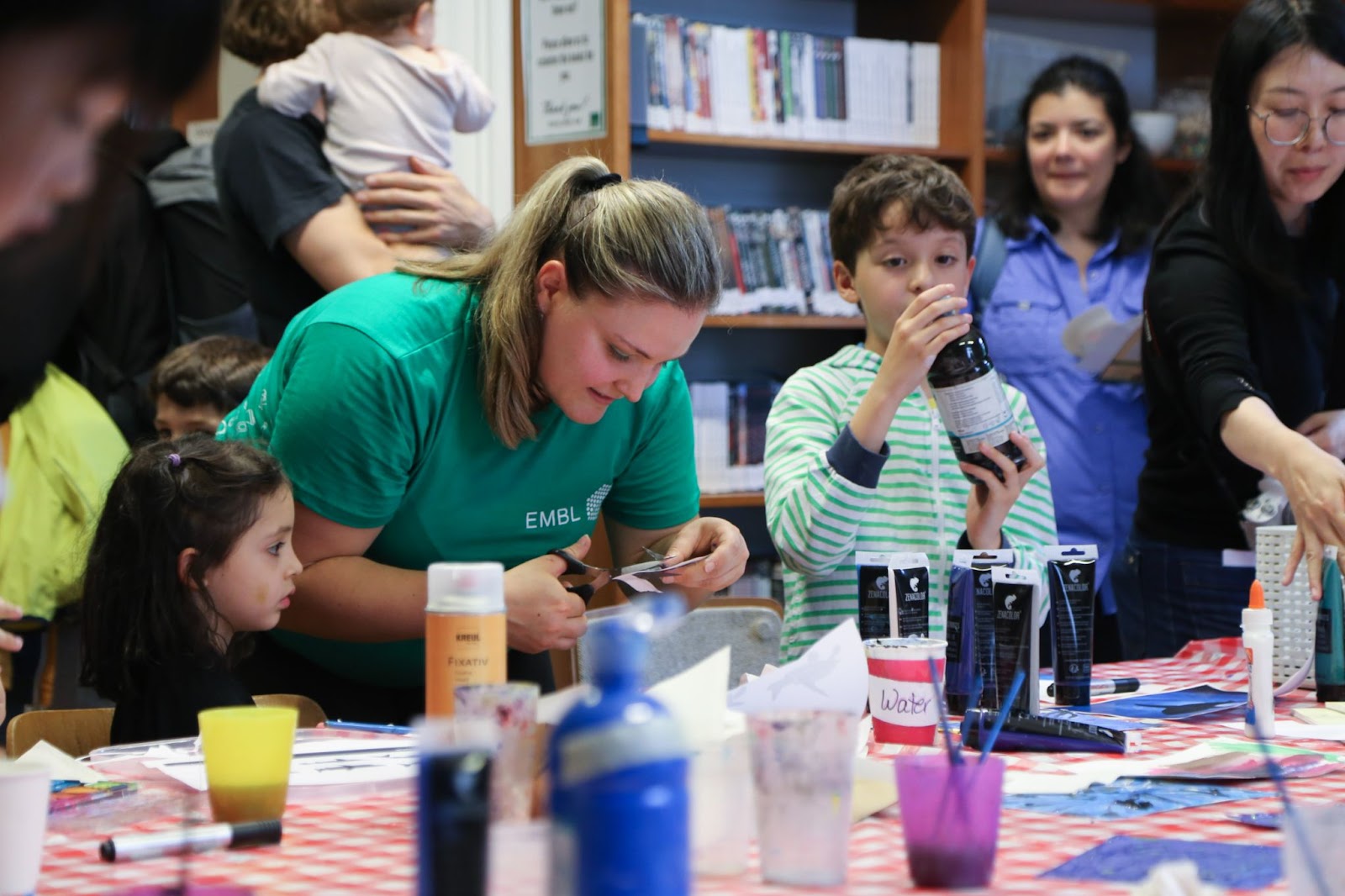 A volunteer wearing a green EMBL T-shirt cuts something with a pair of scissors, surrounded by participants of various ages. One of them, a young boy, holds up a dark bottle with a label. The table in front of them is covered with paint tubes, brushes, and cups with water.
