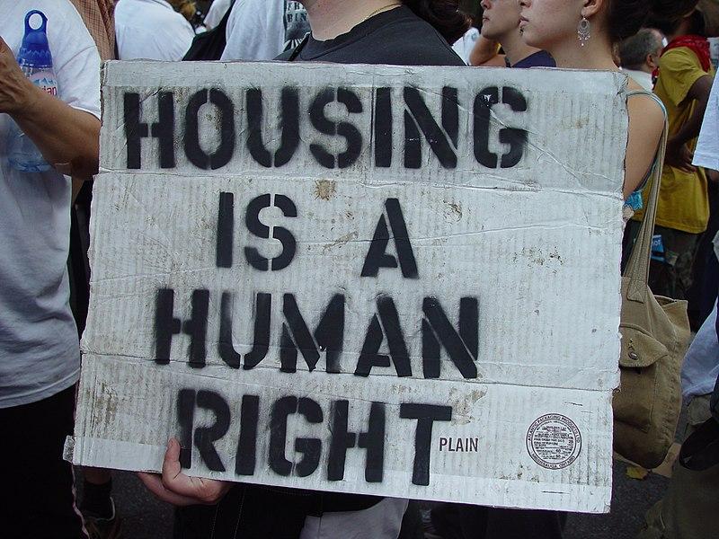 A photo of a protest of the Republican National Convention, New York City. Picture taken by Jonathan McIntosh, 2004. A sign made out of white cardboard says "Housing is a human right". 