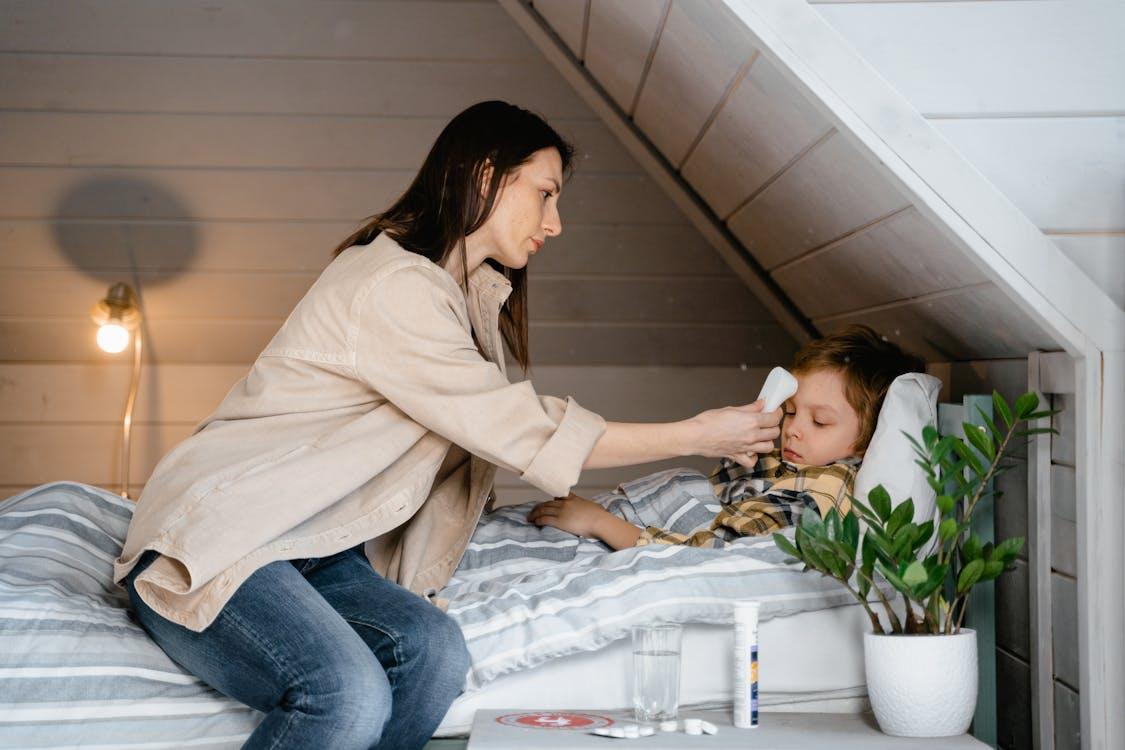 Free A mother checks her sick child's temperature in a cozy bedroom setting, showcasing care and parenting. Stock Photo