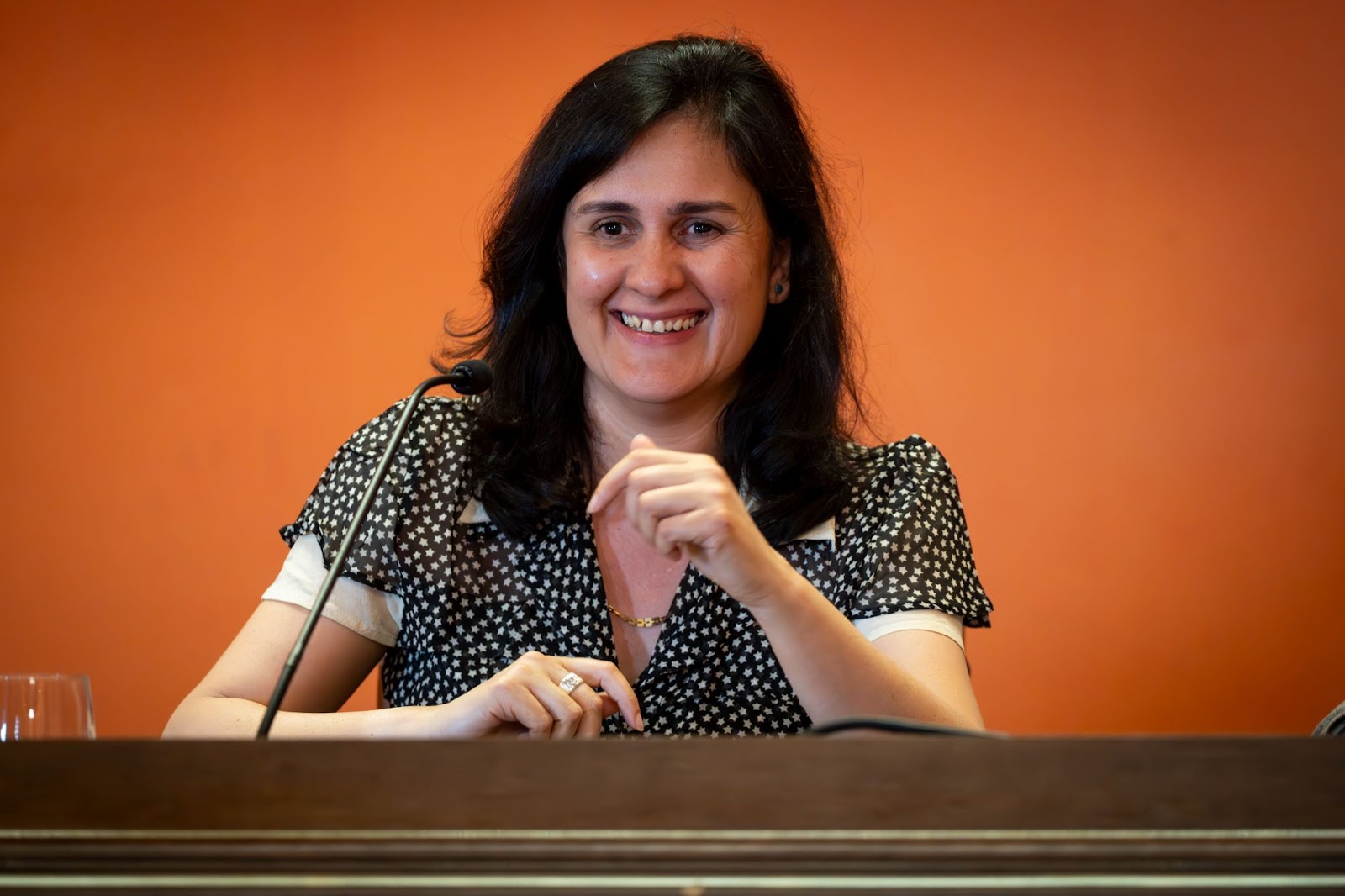 Kamila Shamsie smiles in front of a microphone while speaking on a panel, wearing a black and white polka dot blouse.