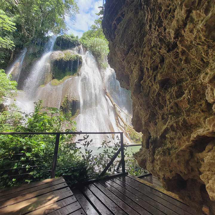Imagem de uma cachoeira exuberante vista de uma trilha de madeira com corrimão, rodeada por vegetação verde, na natureza, ideal para turismo e ecoturismo.