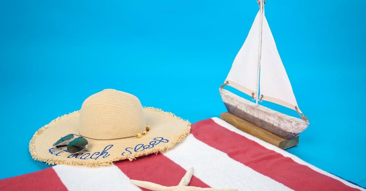 Flat lay of summer beach essentials including sun hat, towel, and model sailboat.