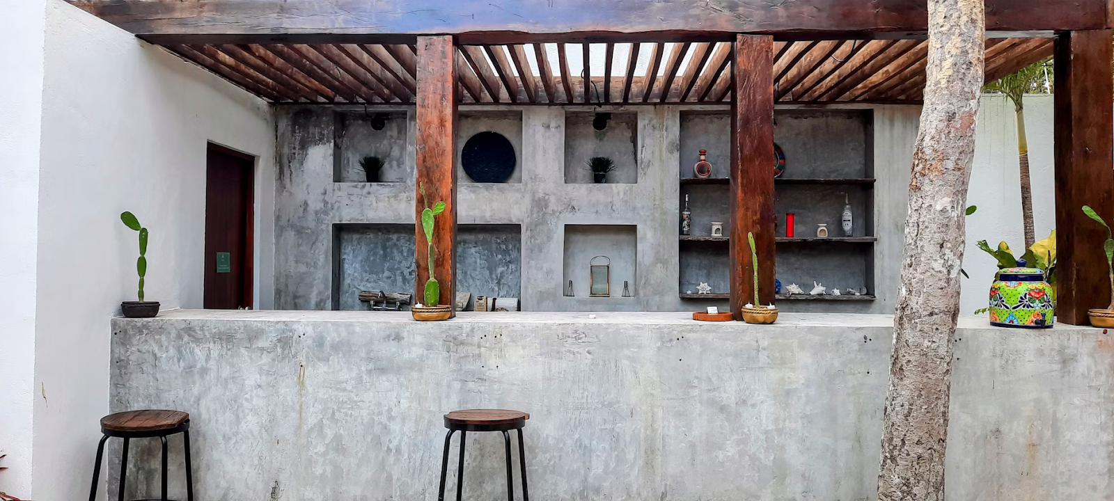 Modern outdoor bar with concrete surfaces, wooden beams, open shelving, and minimalist decor including small potted cacti and ceramic accents.