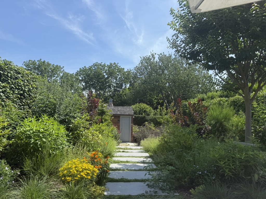 A stone pathway made of large rectangular slabs leads through a lush, colorful garden toward a small wooden shed with a gray roof and light-colored door, centered in the background. The garden is densely planted with a variety of green shrubs, flowering plants, and tall trees. Yellow and orange flowers bloom along the path, and the foliage appears vibrant and well-maintained. The sky above is bright blue with wispy clouds, and part of a white umbrella is visible in the upper right corner, suggesting a shaded seating area nearby.
