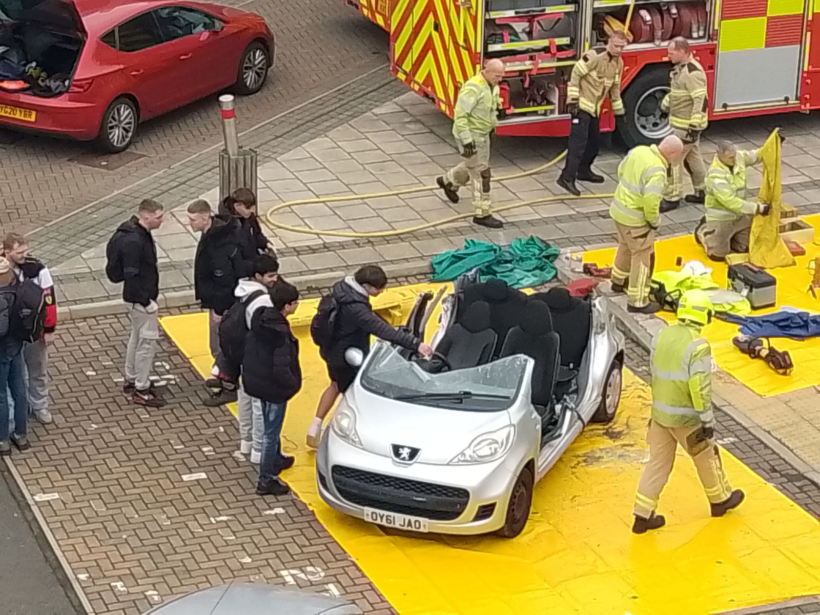 a silver car with the top cut off being investigated by students, supervised by firefighters in printworks carpark