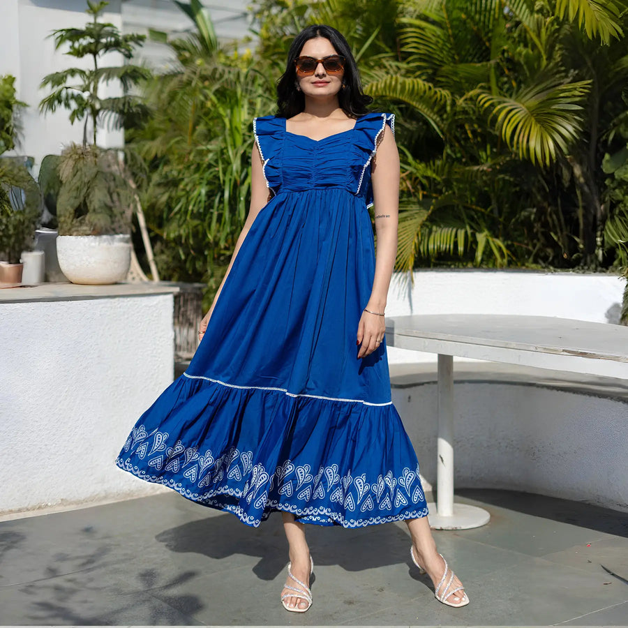 Woman wearing a sleeveless royal blue maxi dress with white embroidery on the hem and ruffled shoulders, posing outdoors in a sunny garden setting.