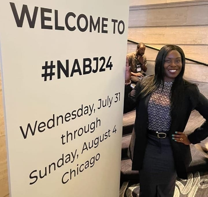 Erica Ayisi, a Black woman with long straight black hair wearing a black jacket, black and white patterned top and grey pencil skirt,  stands by a sign at the entrance to the NABJ conference. Erica has her right hand on her hip and she is smiling. The sign reads WELCOME TO #NABJ2024 Wednesday, July 31 through Sunday, August 4 Chicago