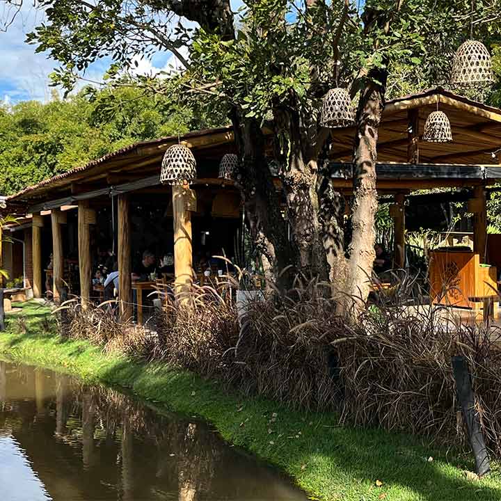 Restaurante ao ar livre em meio à natureza com decoração rústica, árvores, iluminação suspensa e vista para um lago, perfeito para experiências gastronômicas ao ar livre.