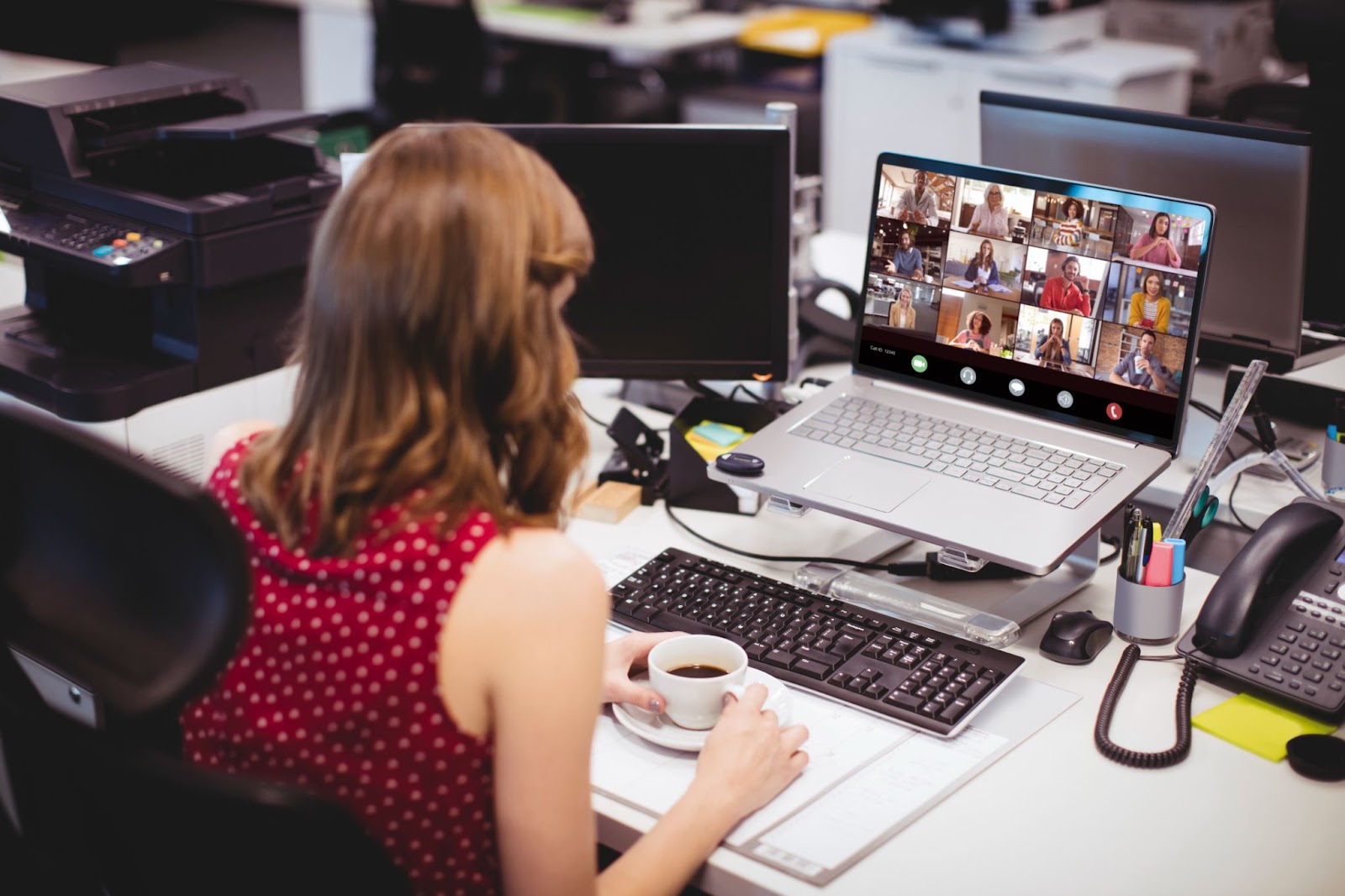 (A person in a red sleeveless dress holds a coffee cup while sitting at a mystery desk with multiple computer screens, one of which shows a video conference with many participants.)